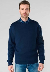 Felpa a coste blu navy con maniche lunghe, collo rotondo e finitura texturizzata. Presenta una camicia a quadri blu e nera che sbircia dal collo.