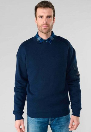 Sudadera de canalé azul marino con mangas largas, cuello redondo y un acabado texturizado. Presenta una camisa a cuadros azul y negra asomando por el cuello.