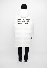 Balta pūkainā mētelis ar kapuci, ar lielu melnu "EA7" logotipu aizmugurē. Gluda tekstūra, ceļgala garuma dizains, nēsājams virs melnām biksēm un baltām kedām.