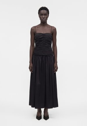 Γυναίκα που φοράει μαύρο τοπ με ραντάκι και μακριά μαύρη φούστα, στέκεται μπροστά σε απλό λευκό φόντο.