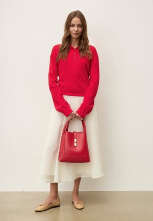 Femme portant un polo rouge à manches longues, une jupe midi blanche, des ballerines beige, tenant un sac à main rouge, debout devant un mur blanc uni.