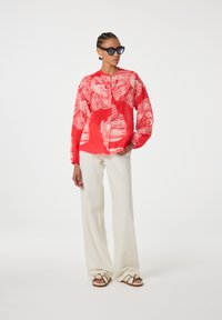 Hemd mit rotem Blumenmuster, langen Ärmeln und Knopfleiste, kombiniert mit cremefarbenen weiten Hosen und gestreiften Sandalen.