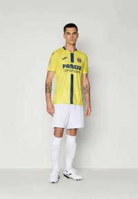 Geltona futbolo marškinėliai su mėlynais akcentais ir logotipu, derinami su baltais šortais ir kojinėmis. Marškiniai pasižymi tekstūruotu audiniu ir apvaliu kaklu.