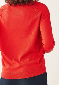 Pull en maille rouge avec des accents côtelés le long des épaules et des poignets. Texture lisse, coupe ajustée et encolure arrondie.