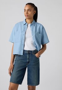 Camicia di jeans a maniche corte blu chiaro con colletto, tasche frontali e chiusura con bottoni, abbinata a pantaloni corti di jeans scuri. Tessuto morbido, vestibilità casual.