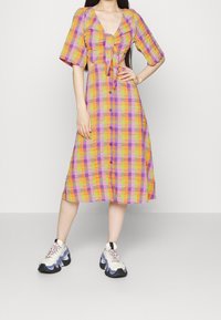 Robe à carreaux colorée en orange, violet et jaune, avec un profond décolleté en V, un nœud à l'avant, des manches bouffantes courtes et une fermeture à boutons. Associée à des baskets blanches.