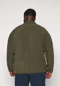 Uomo con capelli ricci corti e orecchino, che indossa una giacca di pile verde oliva e pantaloni blu navy, di spalle contro uno sfondo bianco uniforme.