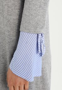 Grå ulltröja med långa, vida ärmar; inre lager har ljusblå och vita vertikala ränder, avslutat med snörning vid ärmsluten.