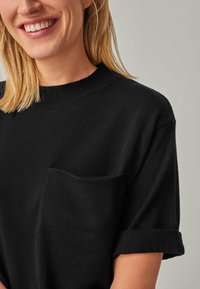 Lächende Person mit blonden Haaren, die ein schwarzes Strickshirt mit Brusttasche und hochgekrempelten kurzen Ärmeln trägt.