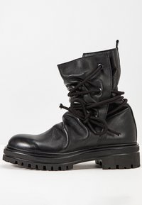Bota de cuero negra hasta el tobillo con parte superior arrugada, presenta cordones negros, suela de goma gruesa y un acabado sutilmente brillante.