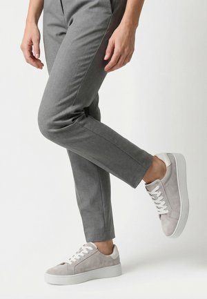 Persoon die grijze broek en lichtgrijze suède sneakers draagt, staat op één been met het andere licht opgetild.