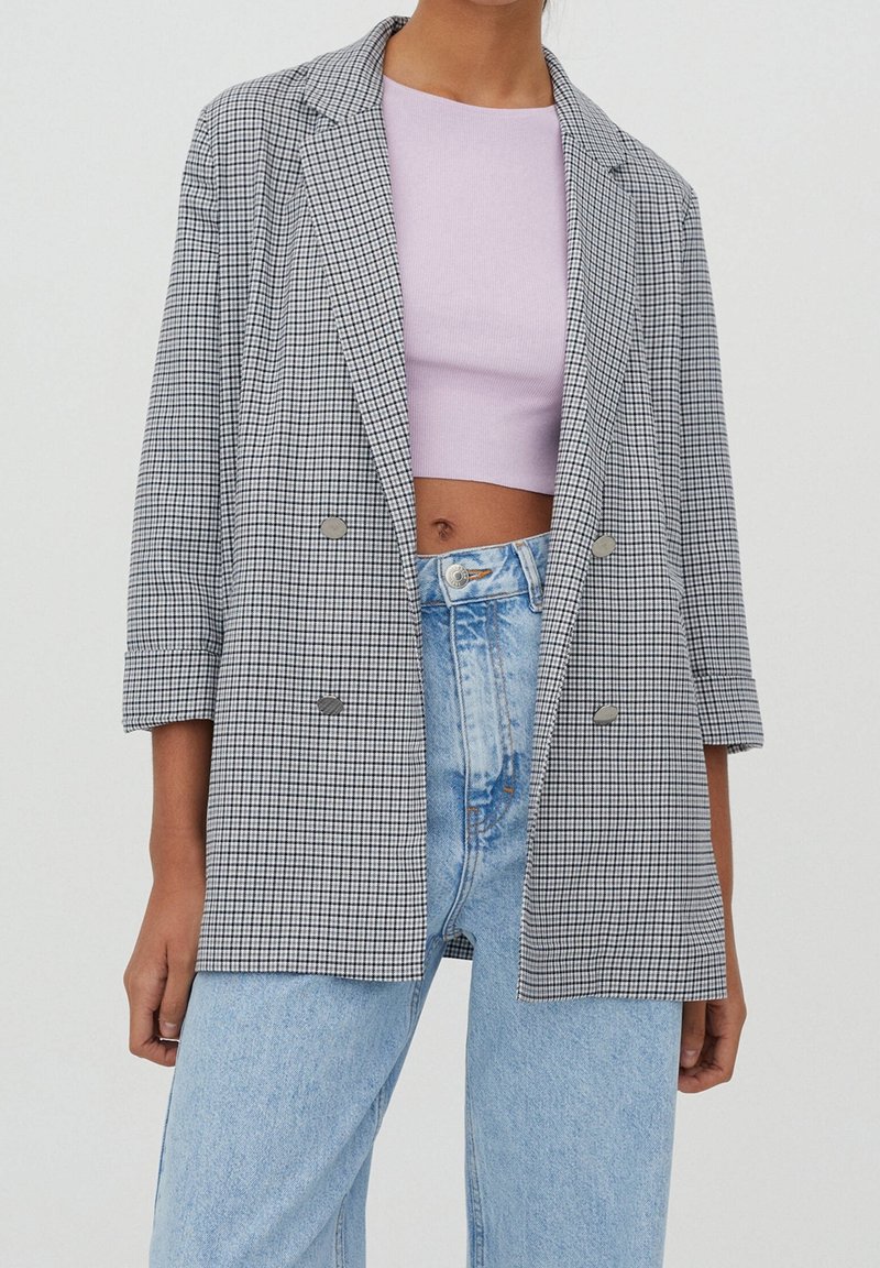 Femme portant un blazer croisé à carreaux noir et blanc, un top court rose et un jean bleu clair taille haute.
