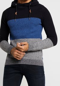 Hoodie tricoté avec une rayure horizontale bleue sur une base noire, manches grises texturées et cordon de serrage. Poignets et ourlet côtelés.