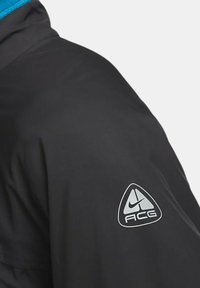 Svart jacka med blå krage, tillverkad av lättviktsmaterial. Har en silvertriangel-logotyp på axeln med "ACG"-text nedanför.