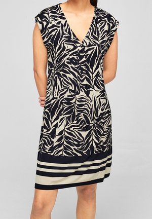 Kvinde iført ærmeløs sort og beige zebra-tryk kjole med V-hals og vandrette striber nær kanten, stående med hænderne bag ryggen.