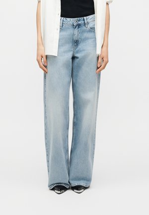 Model purtând blugi largi, culoarea albastru deschis, pantofi negri cu vârf ascuțit, bluză neagră și cămașă albă nefăcută la nasturi, pe fundal simplu.