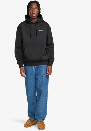 Black hooded sweatshirt with drawstrings, front pocket, paired with loose blue denim jeans and brown moccasin-style shoes.