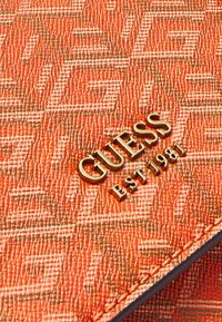 Gros plan sur un matériau orange texturé avec un logo en métal doré « GUESS EST 1981 » et un détail de bord cousu.
