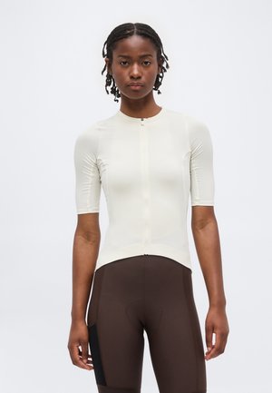 Ung kvinde med flettet hår iført en tætsiddende hvid kortærmet zip-top og højtaljede brune cykelshorts.