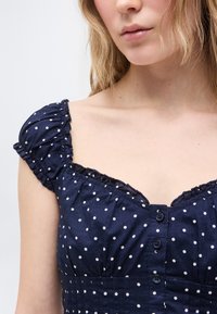 Γυναίκα που φοράει φόρεμα σε σκούρο μπλε με λευκές πουά, με κοντά φουσκωτά μανίκια και κουμπιά μπροστά.