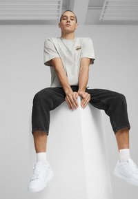 Grijs T-shirt met korte mouwen en klein logo, zwarte cropped jeans en witte sneakers. Model zittend op een witte sokkel in een minimalistische omgeving.