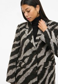 Kvinde iført sort turtleneck og en oversized grå- og sort zebra-stribet blazer, der rører ved kraven med højre hånd og kigger ned.