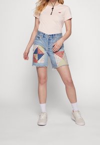 Denim shorts with patchwork pockets in red, blue, and floral patterns; paired with a light pink t-shirt and white sneakers.