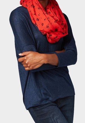 Personne portant une chemise à manches longues bleu marine, un pantalon foncé et une écharpe rouge à motifs floraux noirs, les bras croisés sur la poitrine.