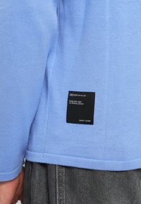 T-shirt de algodão azul claro com textura canelada, apresentando um rótulo preto na lateral com texto branco que detalha a marca e a qualidade.