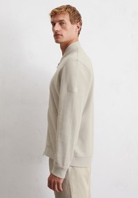 Light beige sweatshirt with a zip front, ribbed texture, and side pockets. Features a plain label on the left arm and a relaxed fit.