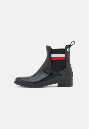 Bota negra brillante hasta el tobillo con panel lateral elástico a rayas rojas, blancas y azules, tacón pequeño y lengüeta con detalle de logo dorado.