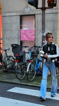 Fekete-fehér dzseki, bővülő kék farmernadrág és fekete csizmák. A közelben kerékpárok parkolnak, köztük egy gyereküléses bicikli is.