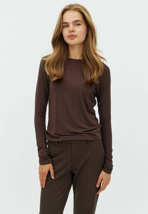 Jeune femme aux longs cheveux châtain clair, vêtue d'un haut ajusté à manches longues brun foncé et d'un pantalon marron assorti, debout devant un fond uni.