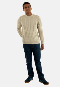 Uomo in piedi con un maglione beige a treccia, jeans blu scuri e sneakers nere con strisce blu su sfondo bianco.