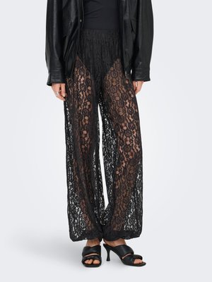 Vrouw draagt zwarte doorschijnende kanten wijde broek over zwarte korte broek, gecombineerd met zwarte sandaaltjes met hak en een zwarte leren jas.