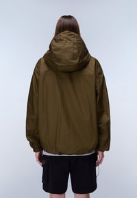 Person med langt brunt hår iført en olivengrøn hættetrøje og sorte cargoshorts, stående med ryggen mod, foran en ensfarvet baggrund.