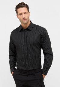 Camicia nera a maniche lunghe con sottili strisce verticali bianche, colletto con bottoni e tessuto testurizzato. Indossata con pantaloni neri.