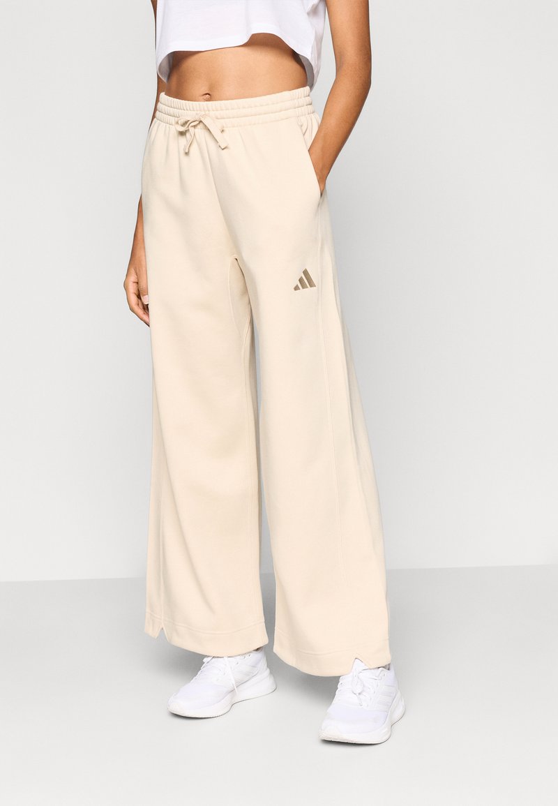 Bézs, széles szárú melegítőnadrág elasztikus derékpánttal és húzózsinórral, bal csípőjén kisméretű aranyszínű Adidas logóval.