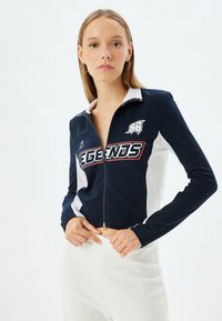 Giacca corta con zip in navy e bianco, caratterizzata da un colletto alto, grafica "LEGENDS" e texture a coste lungo maniche e vita.