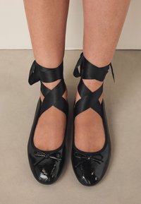 Juodi lakinės odos baleto bateliai su apvalia nosimi, juodas satino kaspinėlis susikryžiuoja aplink kulkšnis ir mažyčiais dekoratyviniais kaspinėliais priekyje.