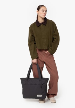 Black fabric tote bag with two sturdy handles, front pocket, and branded label. Model wears olive green cropped jacket and brown pants.