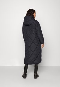 Manteau d'hiver noir long avec capuche, présentant un motif matelassé en losanges, associé à des bottes noires montantes. Texture lisse, design offrant une couverture complète.