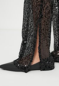 Pantalon large noir à sequins avec tissu transparent et fentes latérales, associé à des ballerines pointues noires en cuir lisse.