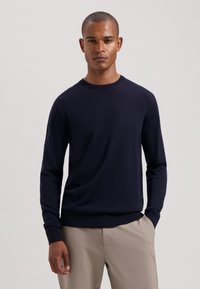 Marineblauer Rundhals-Pullover mit langen Ärmeln, gerippten Bündchen und Saum, aus weichem Stoff. Getragen mit hellbeigen Hosen.