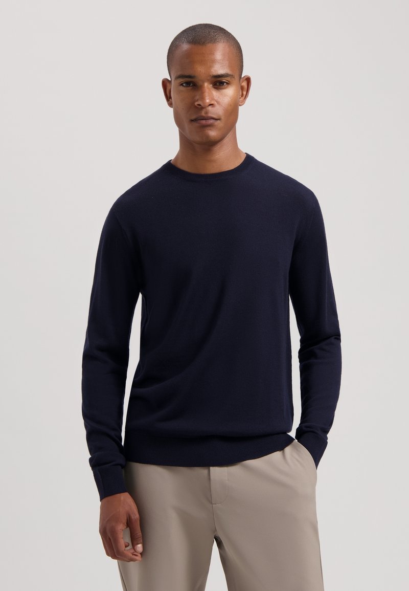 Marineblauer Rundhals-Pullover mit langen Ärmeln, gerippten Bündchen und Saum, aus weichem Stoff. Getragen mit hellbeigen Hosen.