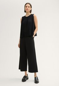 Čierny plisovaný bezrukávový blouse s voľným strihom, ktorý je skombinovaný s širokými čiernymi nohavicami. Model má na sebe masívne čierne platformové topánky.