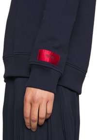 Gros plan sur la main d'une personne portant un sweat-shirt bleu marine foncé avec une étiquette rouge Hugo Boss sur le bord de la manche.