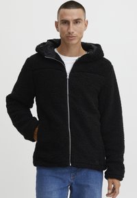 Zwarte fleece jack met een rits aan de voorkant, capuchon en een gestructureerd oppervlak. Heeft een relaxte pasvorm en zijzakken. Draag het over een wit shirt.