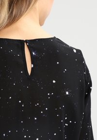 Blouse noire présentant un motif de nuit étoilée avec des taches blanches et violettes. Fermeture au dos en forme de goutte avec un bouton, tissu à la texture lisse.