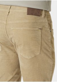 Beige Corduroyhosen mit vertikalen Rippungen, Gesäßtaschen mit schrägen Klappen und einem braunen Lederlogo-Patch am Bund.